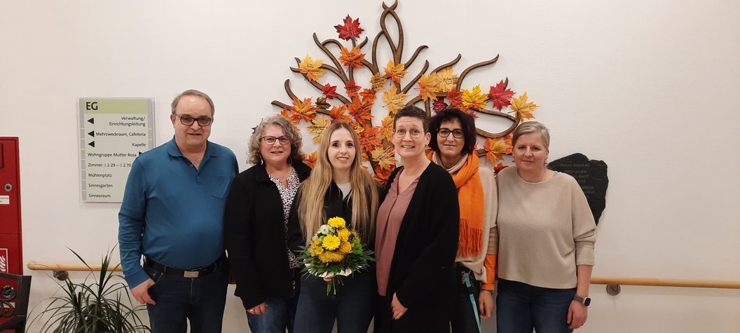 Sechs Personen stehen vor einer Wand mit herbstlicher Dekoration aus bunten Blättern, eine Frau in der Mitte hält einen Blumenstrauß.