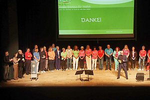 Gruppe von Menschen steht auf einer Bühne vor einer grünen Leinwand mit dem Text 'DANKE!'