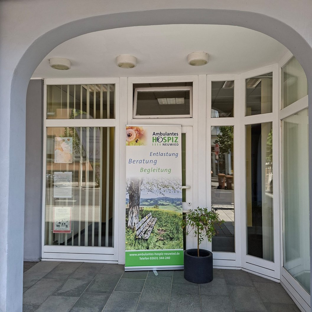 Eingangsbereich mit weißem Bogen, Glastür und seitlichen Fenstern, davor ein Roll-up mit Text und Bild eines Baums und einer Landschaft