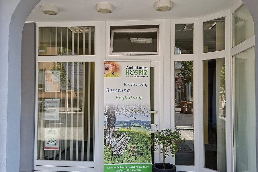 Eingangsbereich mit weißem Bogen, Glastür und seitlichen Fenstern, davor ein Roll-up mit Text und Bild eines Baums und einer Landschaft