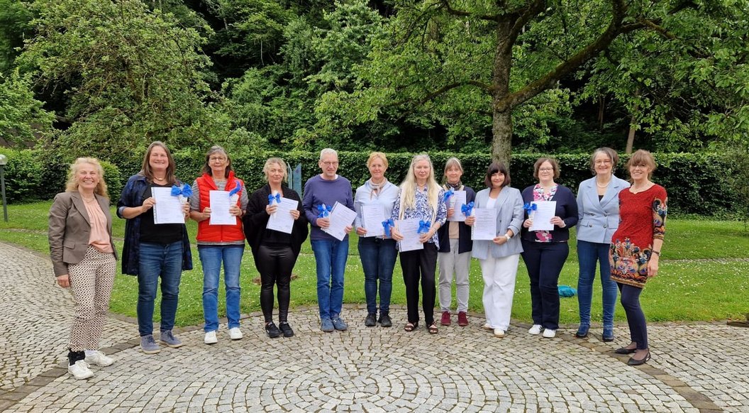 Gruppe von elf Personen steht auf gepflastertem Platz vor Bäumen, einige halten Papiere in der Hand.