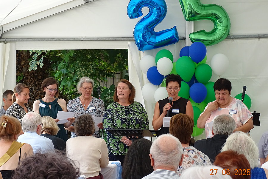 Gruppe von Menschen sitzt und steht in einem Zelt vor Publikum mit blauen und grünen Luftballons in Form der Zahl 25