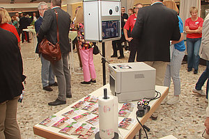 Tisch mit Fotobox, Drucker und Fotostreifen, im Hintergrund Menschen in Gesprächsrunden auf einer Veranstaltung