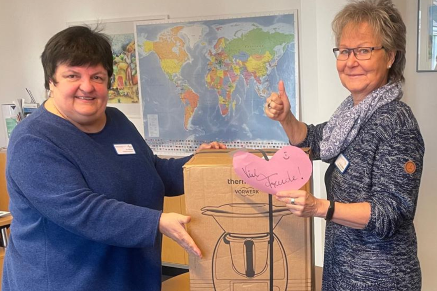 Zwei Frauen stehen in einem Raum mit Weltkarte im Hintergrund und halten gemeinsam einen großen Karton mit einer Küchenmaschine