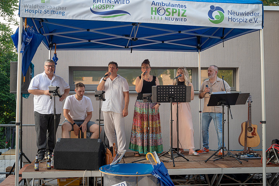 Sechs Personen auf einer Bühne unter einem blauen Zelt mit Logos von Hospiz-Organisationen, einige singen oder sprechen in Mikrofone, eine Person spielt Cajón.