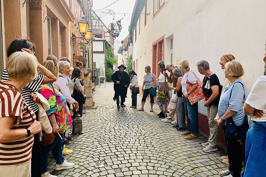 Menschen stehen in einer engen gepflasterten Gasse in zwei Reihen gegenüber und hören einer Person in historischer Kleidung zu