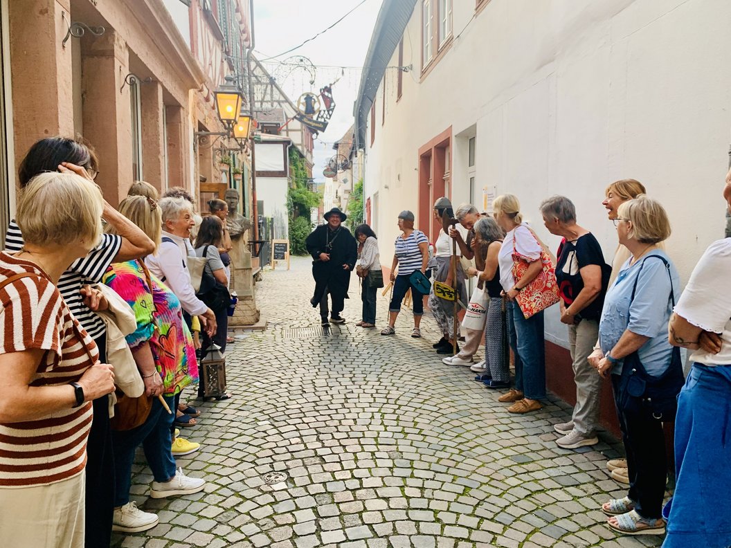 Menschen stehen in einer engen gepflasterten Gasse in zwei Reihen gegenüber und hören einer Person in historischer Kleidung zu