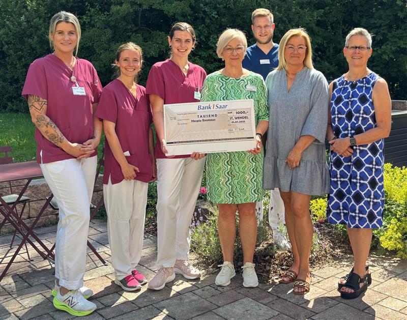 Sechs Frauen stehen im Freien auf einem gepflasterten Weg, zwei tragen bordeauxrote Pflegekleidung, eine hält einen großen Scheck mit der Aufschrift 'Bank1Saar'