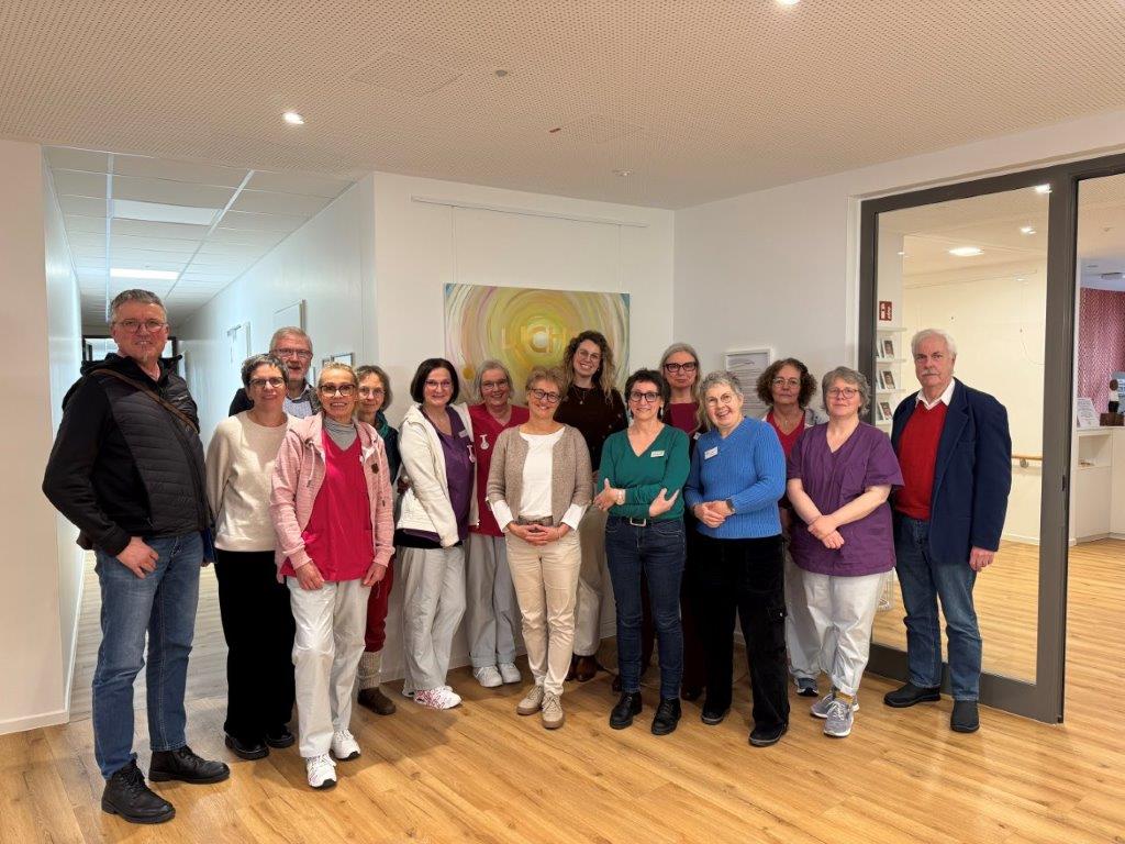 Gruppe von 13 Personen steht in einem hellen Raum mit Holzboden und weißer Wand, einige tragen bunte Kleidung, im Hintergrund ein abstraktes Gemälde.