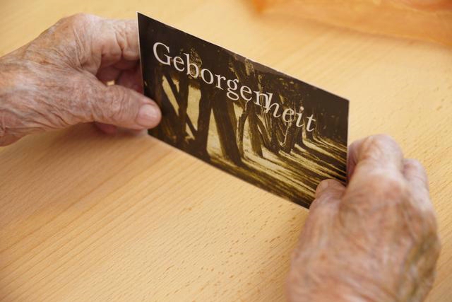 Zwei ältere Hände halten eine Karte mit dem Wort 'Geborgenheit' auf einem Holztisch