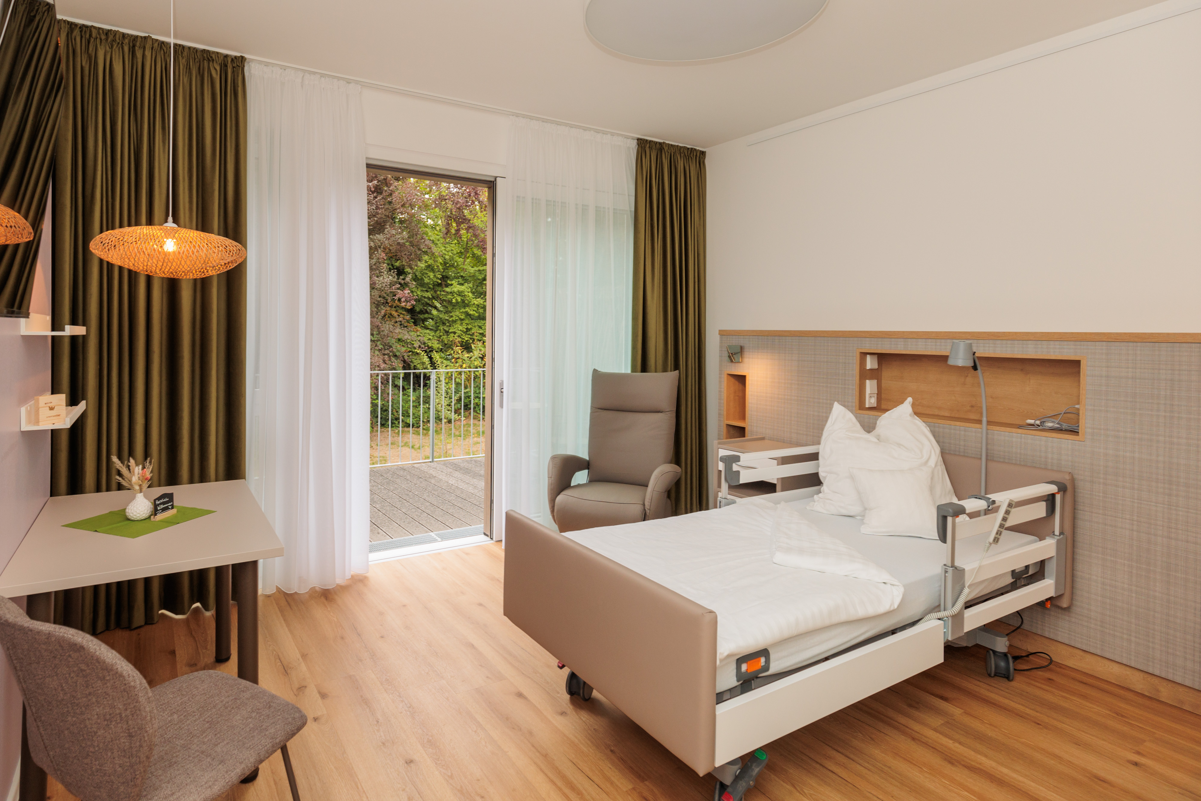 Modernes Patientenzimmer mit Pflegebett, Sessel, Tisch mit Stuhl und Balkon mit Blick ins Grüne
