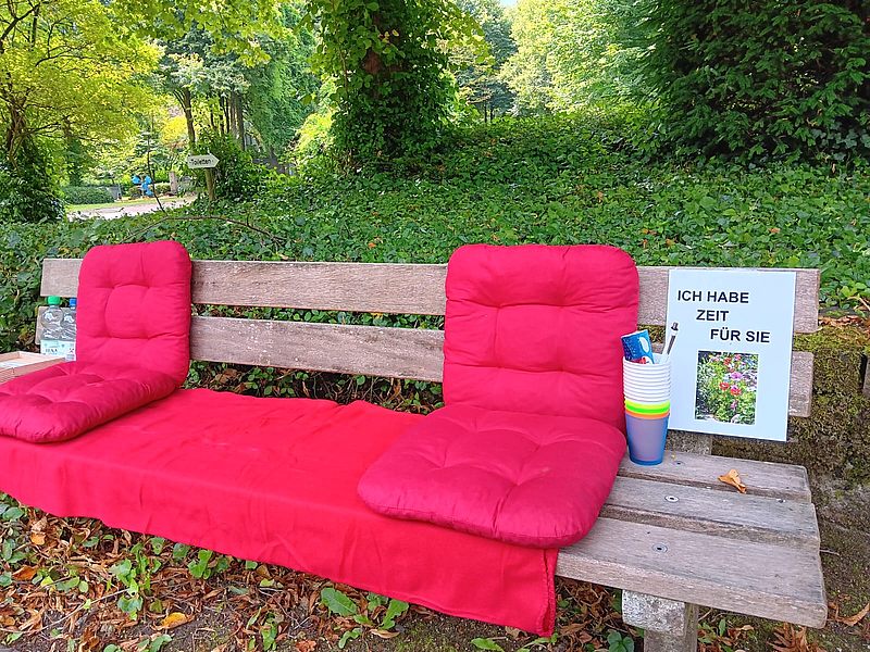 Holzbank im Park mit roten Sitzkissen und einem Schild mit der Aufschrift 'Ich habe Zeit für Sie' neben bunten Bechern